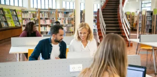 Międzynarodowa grupa studentów pracuje przy laptopach w bibliotece kampusu.