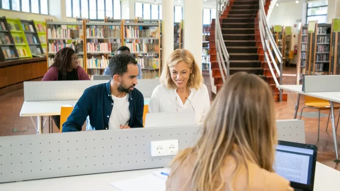 Międzynarodowa grupa studentów pracuje przy laptopach w bibliotece kampusu.